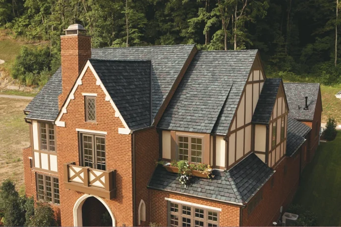 Grand Manor Colonial Slate by CertainTeed on a home. The roof is a dark gray slate, and the house is brick with Tudor-style accents. Lush green trees behind.