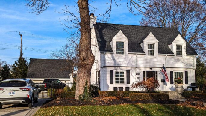 White two-story home with black shutters and matching roof shingles featuring dormer windows and an American flag displayed near the front entrance