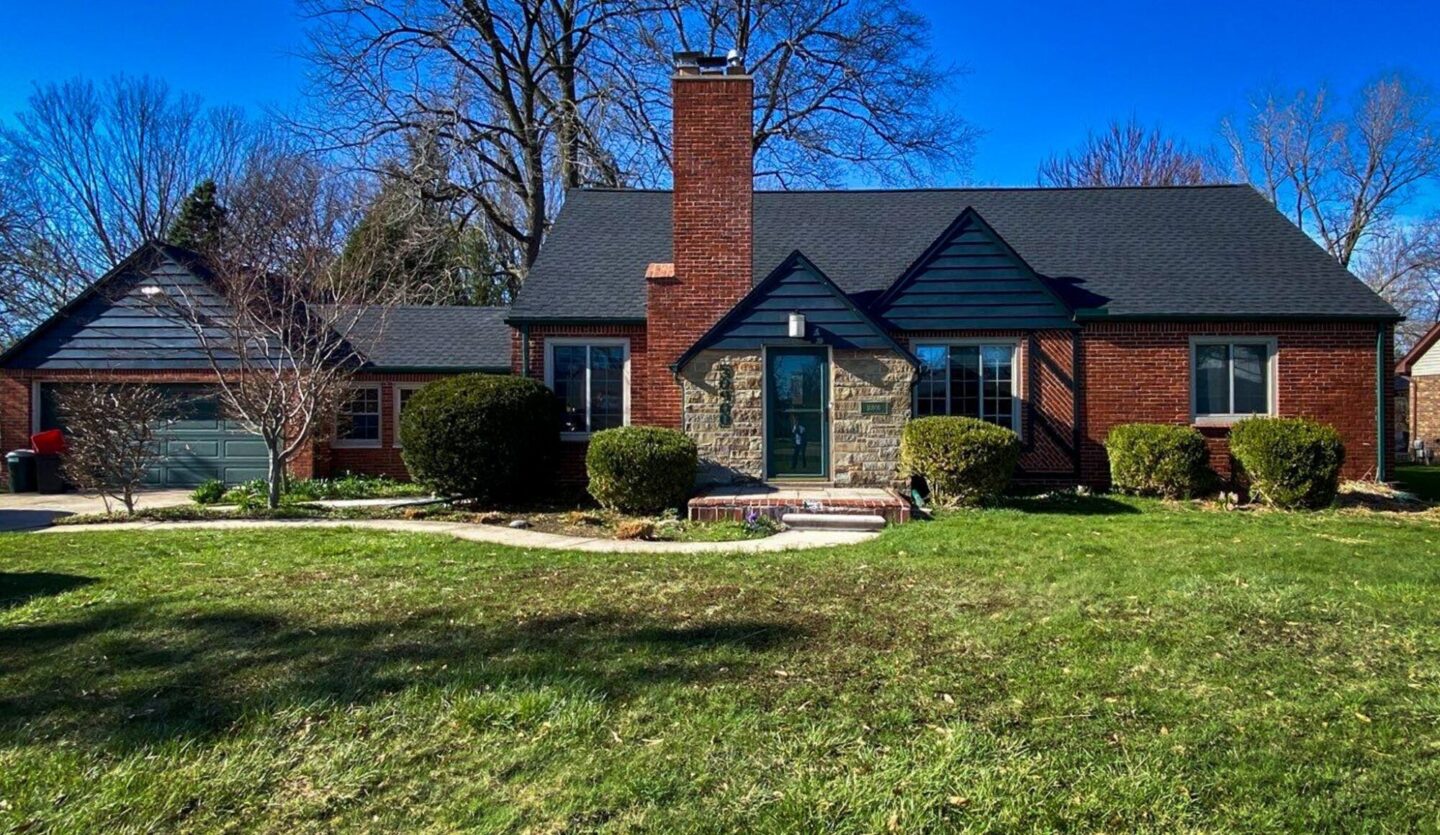 Livonia Rayburn roof replacement. Front view of a brick house with a new roof. Landscaped lawn and blue sky on a sunny day.