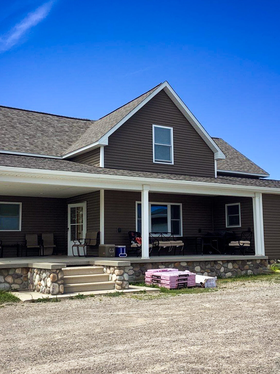 Posen roof replacement. Front view of a brown house with a porch and a brown roof. The house is surrounded by a gravel driveway and a blue sky.