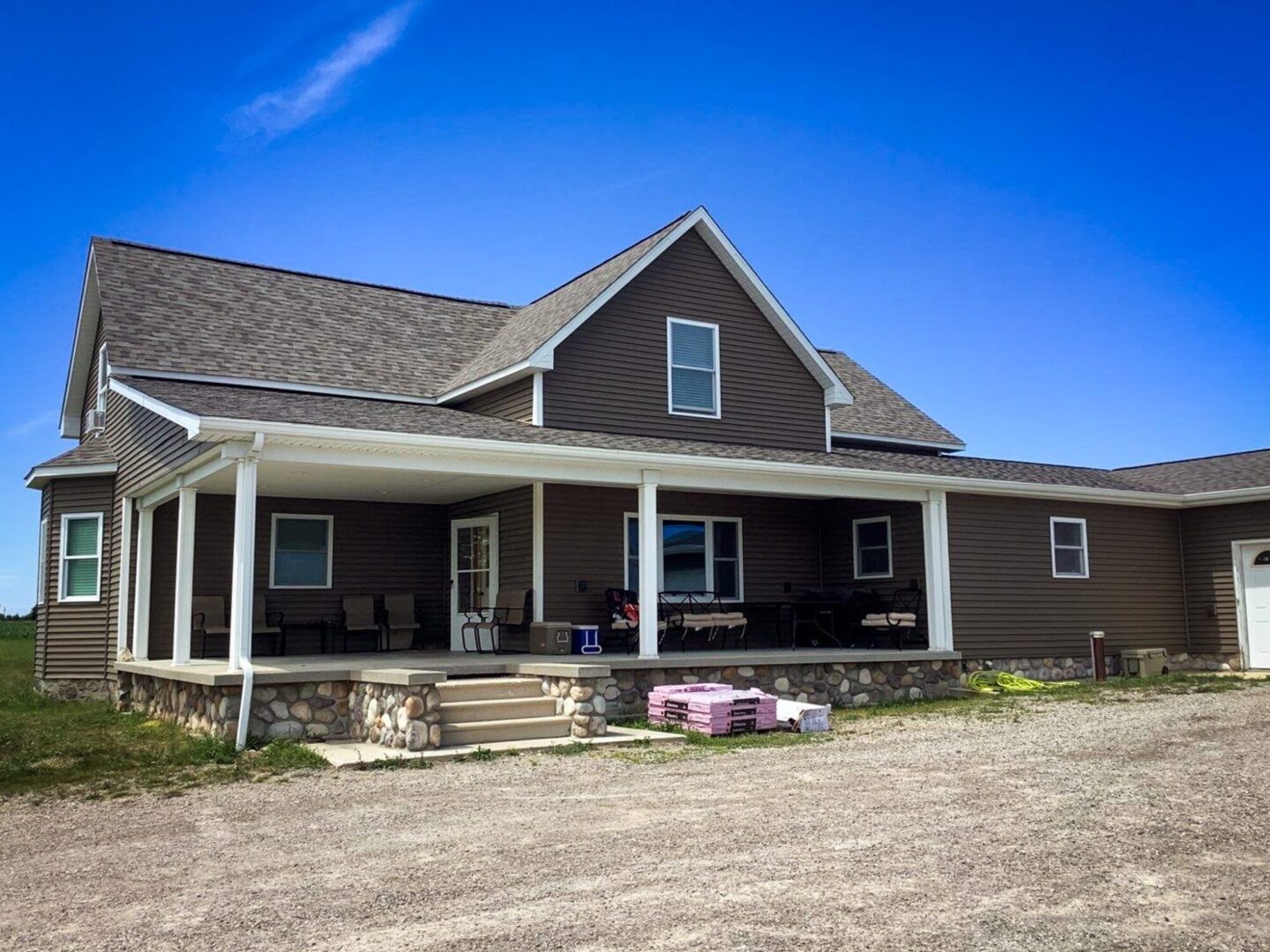 Posen roof replacement. Front view of a brown house with a porch and a brown roof. The house is surrounded by a gravel driveway and a blue sky.