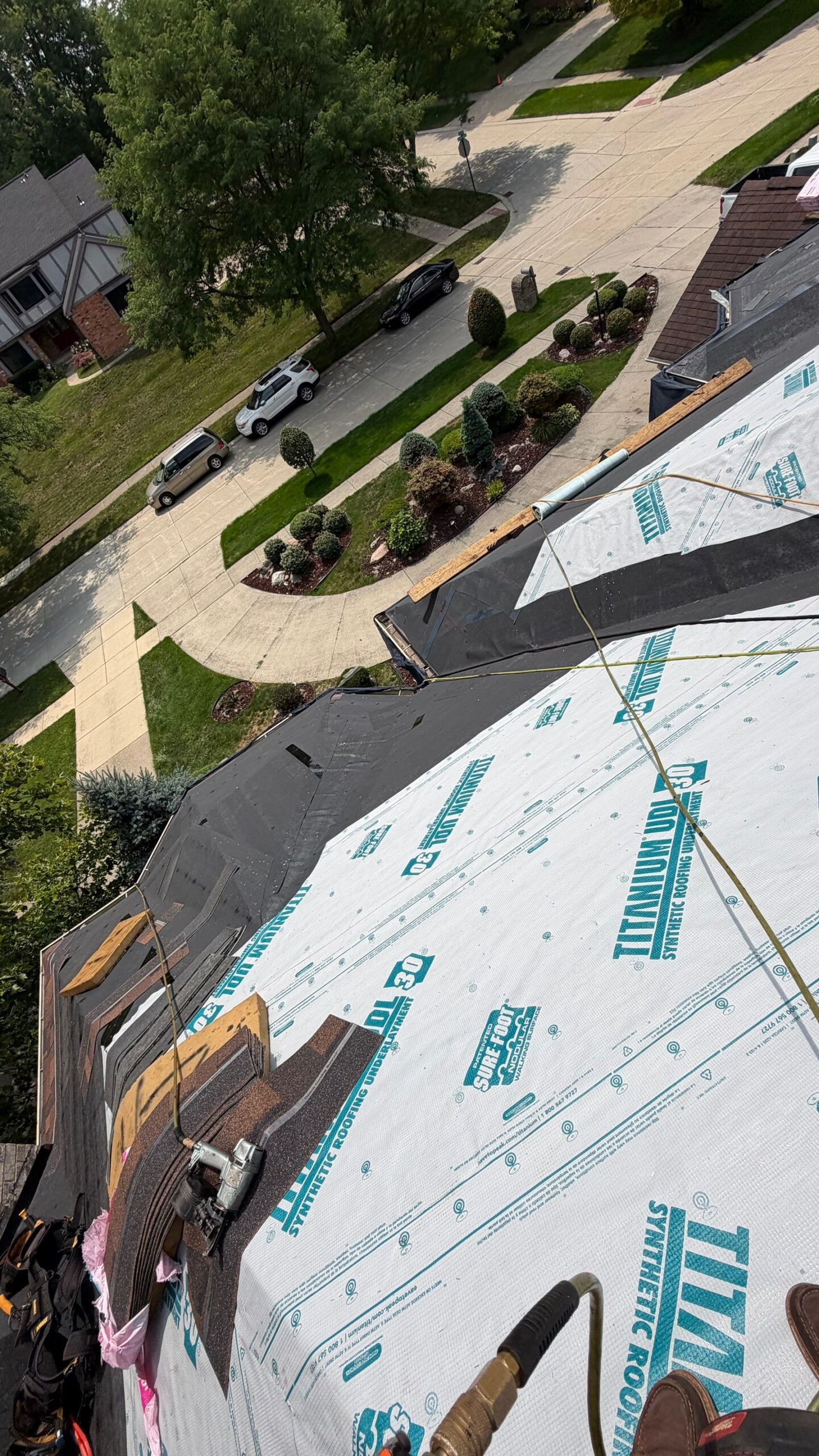 Roof under construction with synthetic underlayment, exposed shingles, tools, and a residential neighborhood visible below