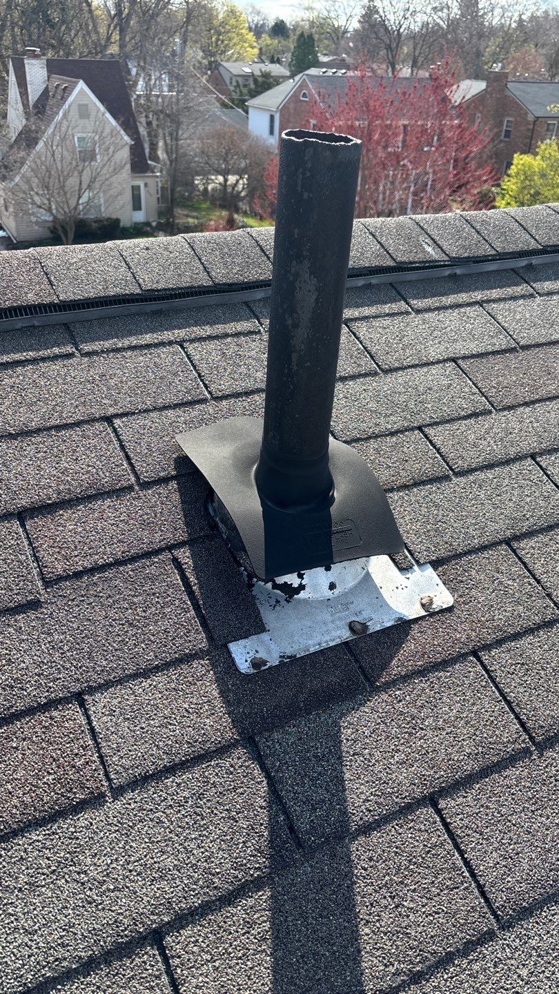 Black roof vent pipe extending through asphalt shingles, with houses and leafless trees in the background