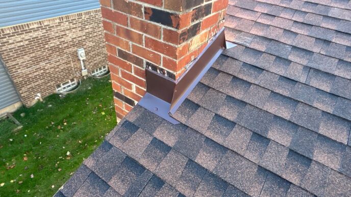 Close-up view of new asphalt shingles installed around a red brick chimney on a residential roof in Livonia featuring clean metal flashing perimeter work.