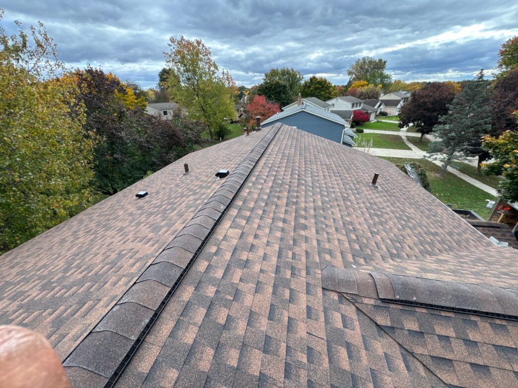 Completed brown multi-tonal asphalt shingle roof replacement on a suburban home in Livonia showing ridge vents and plumbing pipes under a cloudy sky.