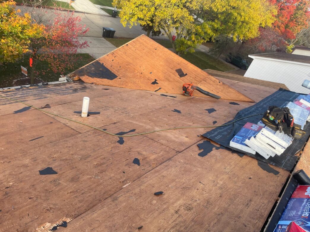 Exposed plywood roof decking during a Livonia home replacement project showing a power tool sitting on the sloped surface near a white plumbing vent pipe.
