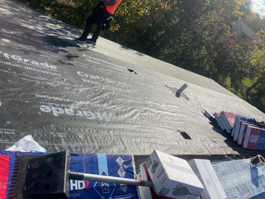 Worker standing on a sloped roof covered in black synthetic underlayment featuring CraftGrade branding, with stacks of GAF shingles and tools nearby.