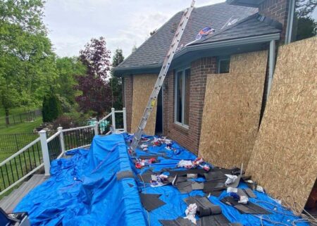 New Baltimore roof replacement process showing a house with a ladder against the roof, covered in a blue tarp, with old shingles removed and debris scattered around.