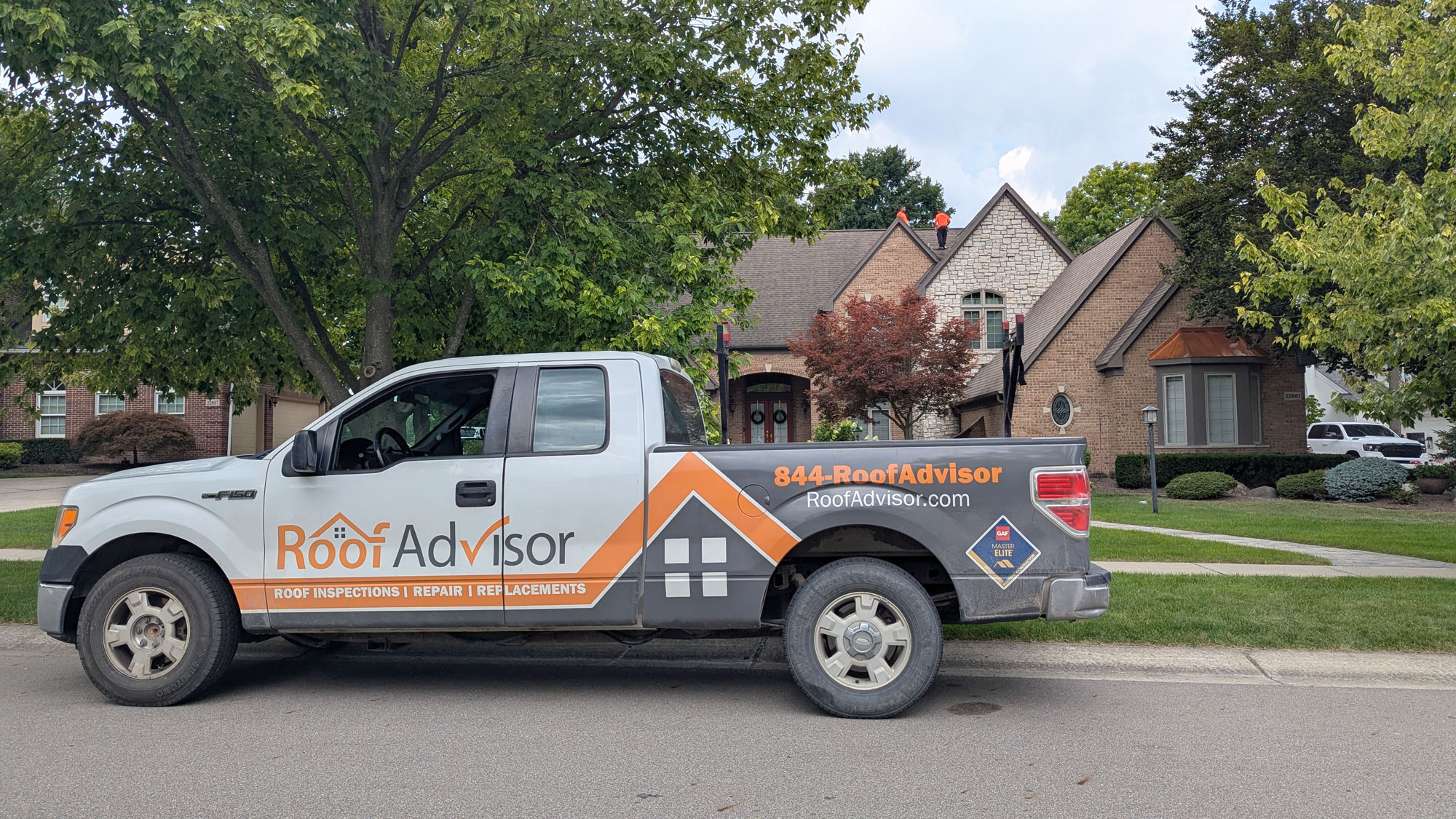 RoofAdvisor truck on-site for roof inspections, repair, and replacements. The company truck is parked in front of a house with workers on the roof.